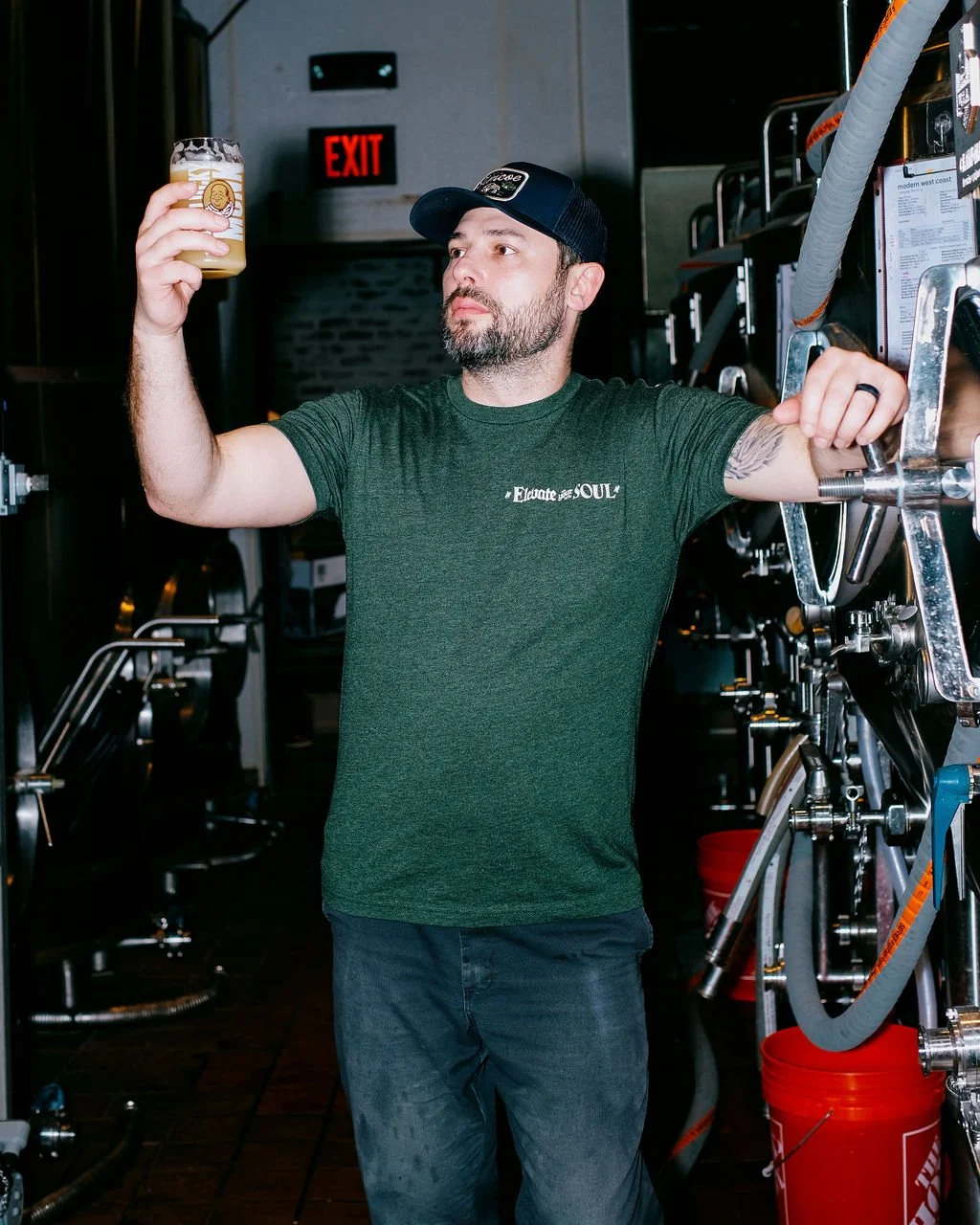 A man with a beard in a green t-shirt and black cap holding a glass of beer, standing next to brewing equipment in a brewery or taproom.