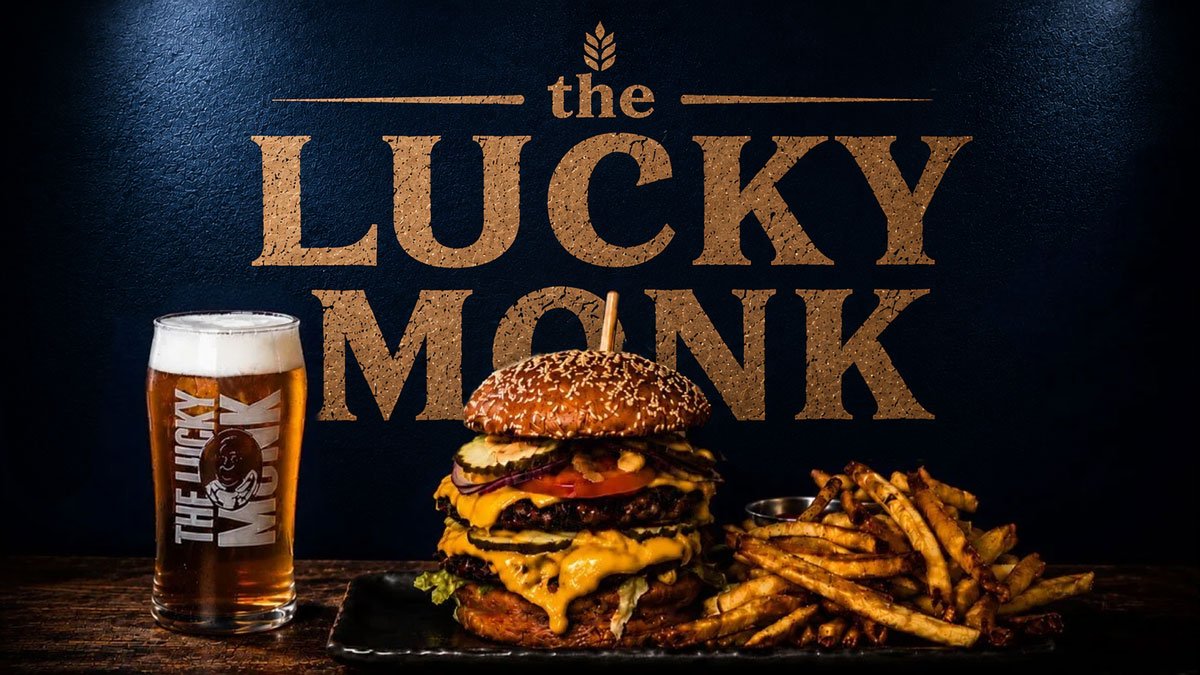 A tall glass of beer, a large double cheeseburger with multiple patties, cheese, pickles, lettuce, tomato, onions, and a sesame seed bun, along with a side of French fries, in front of a sign that says "the LUCKY MONK."
