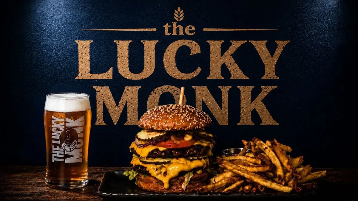 A large burger with multiple beef patties, cheese, pickles, tomato, lettuce, and a sesame seed bun, served with a side of French fries and a glass of beer, against a dark background with the text "the lucky monk."