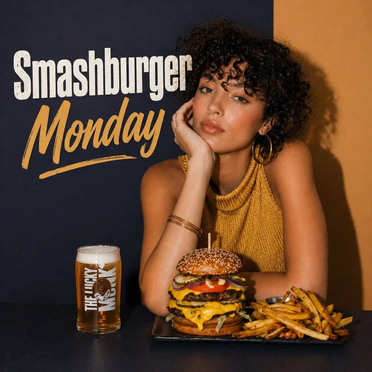 A woman with curly hair and gold jewelry sitting at a table with a large cheeseburger, fries, and a glass of beer, with a sign behind her that reads 'Smashburger Monday.'