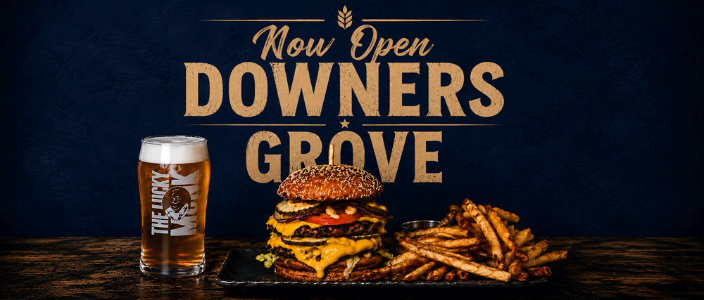 A large burger with multiple patties, cheese, lettuce, tomato, and toppings sits on a black plate. Next to it are a pile of French fries. To the left is a tall glass of beer with a logo that reads 'The Lucky Monkey.' Behind, there is a sign that says 'Now Open the Downers Grove' in bold letters on a blue wall.
