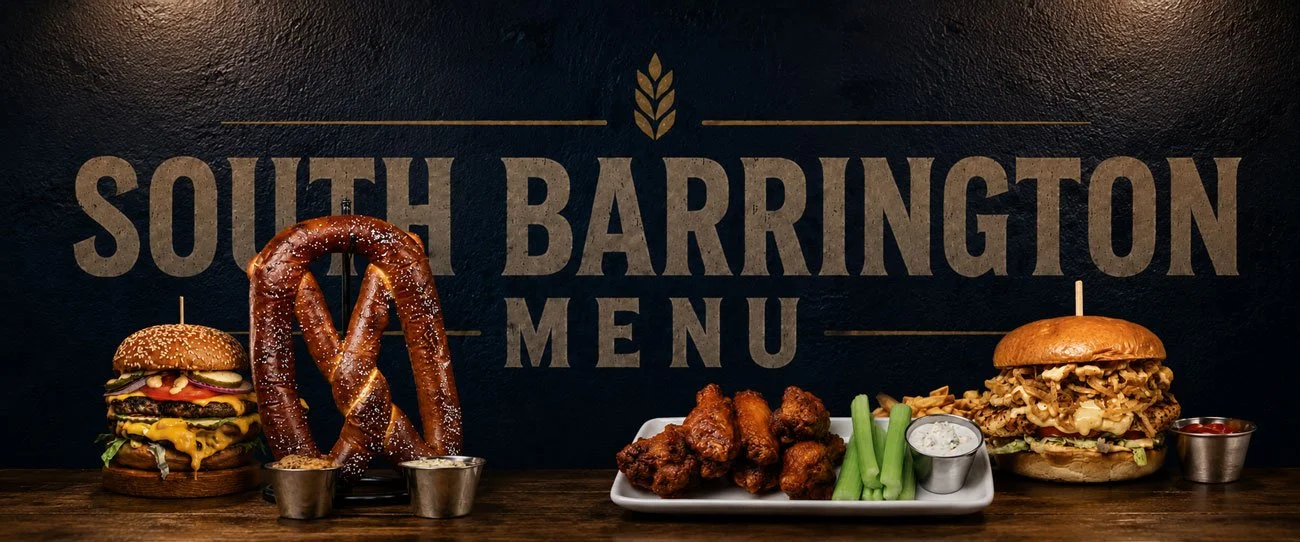 A menu board with the text 'South Barrington Menu' in the background, featuring a large pretzel, two cheeseburgers with toppings, chicken wings with celery and ranch, and small containers of dipping sauces on a wooden table.