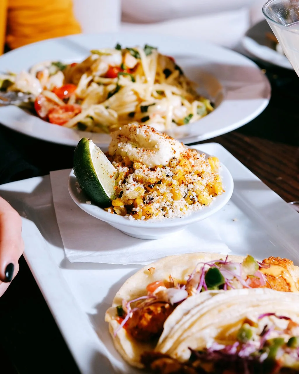A plate of Mexican street food including a taco filled with meat and toppings, a bowl of Mexican street corn with cheese and lime, a plate of pasta with vegetables, and a bowl of ceviche with lemon.
