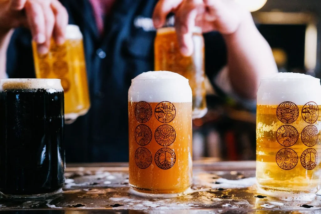 Various glasses of beer, including a dark stout and lighter ales, on a wet bar counter with a bartender pouring beer in the background.