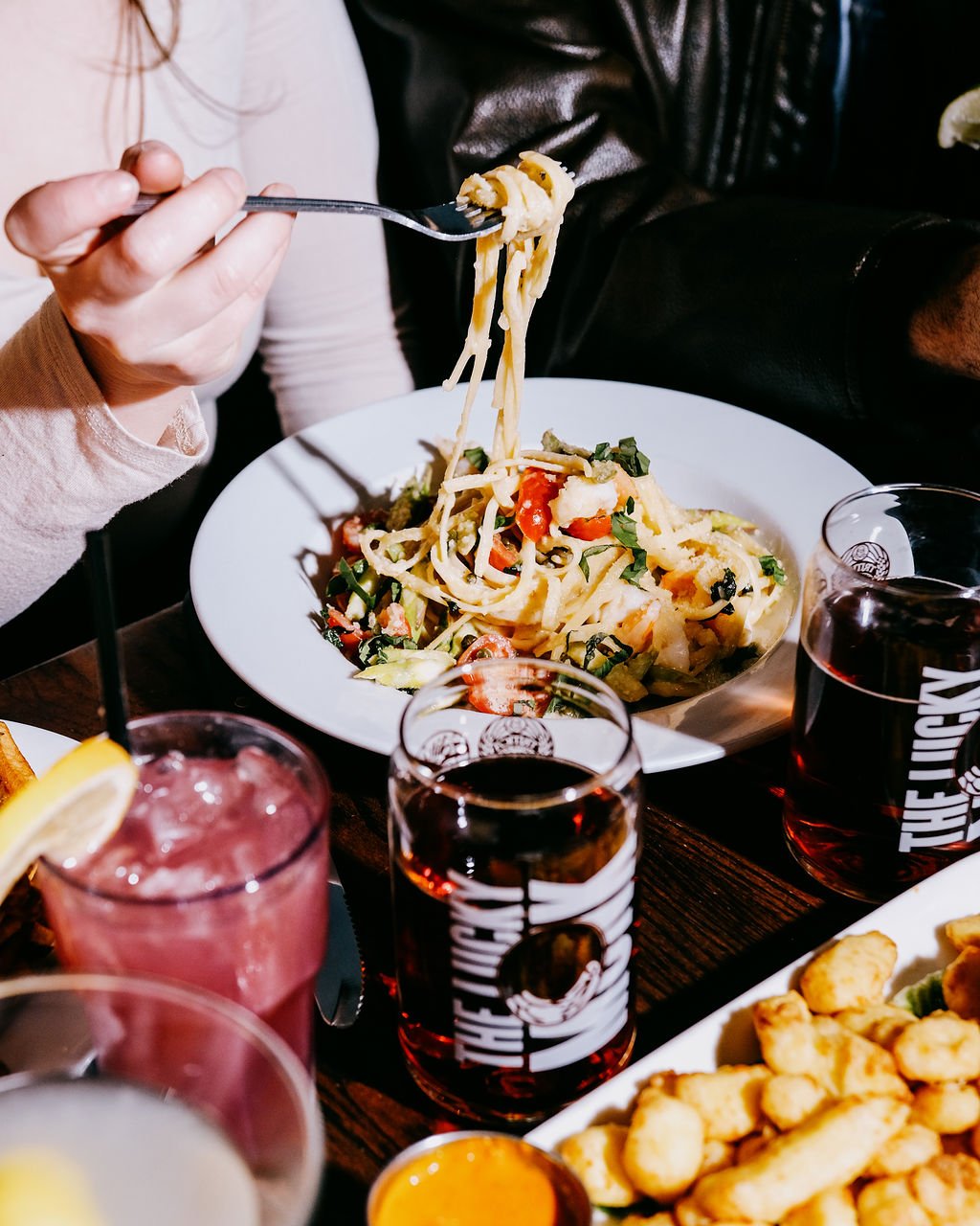 Person lifting forkful of pasta with vegetables at a dining table with drinks and other dishes.