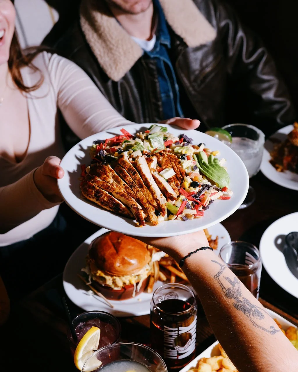 People sharing a meal with a chef presenting a salad with grilled chicken.