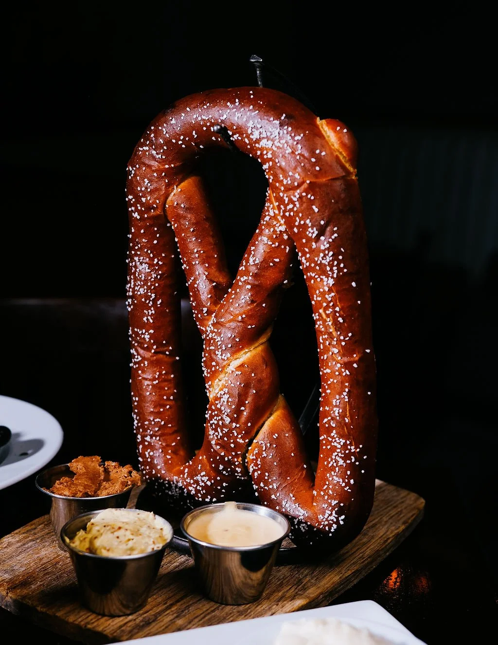 Large salted pretzel on a wooden board with three small metal cups of dips and condiments.