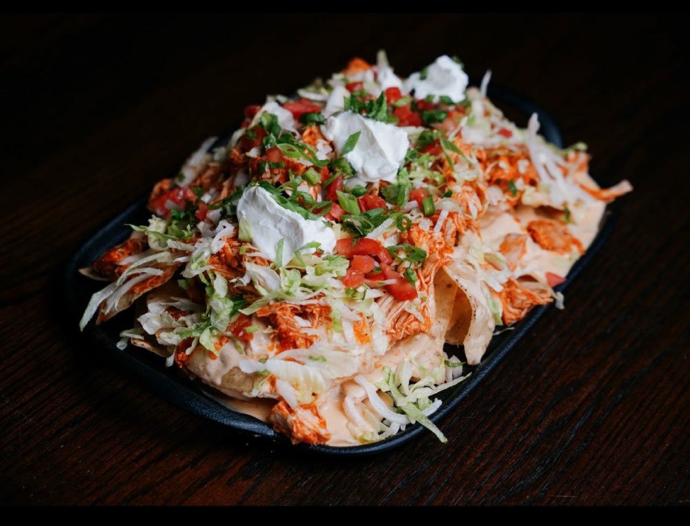 Plate of loaded nachos topped with shredded cheese, chopped tomatoes, green onions, and dollops of sour cream, on a dark wooden table.