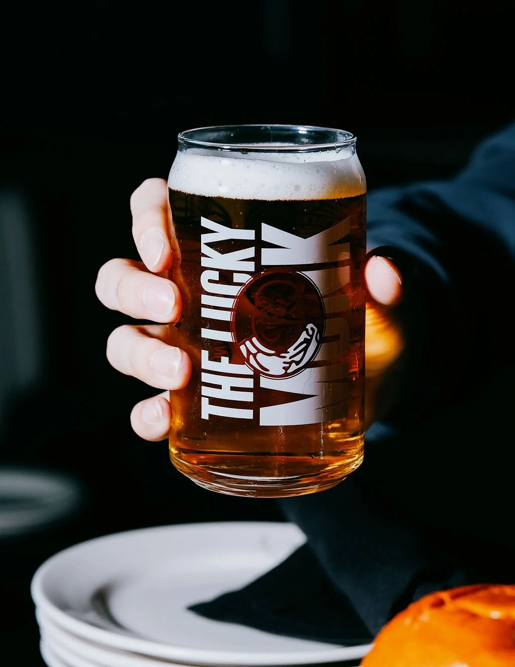 A person holding a glass of beer with the logo 'The Lucky Wolf' on it, in a dark setting.