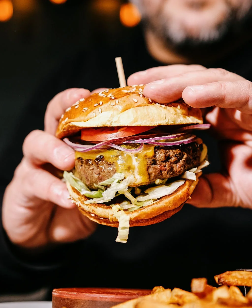 Close-up of a person holding a cheeseburger with lettuce, tomato, red onion, cheddar cheese, and a beef patty in a sesame seed bun.