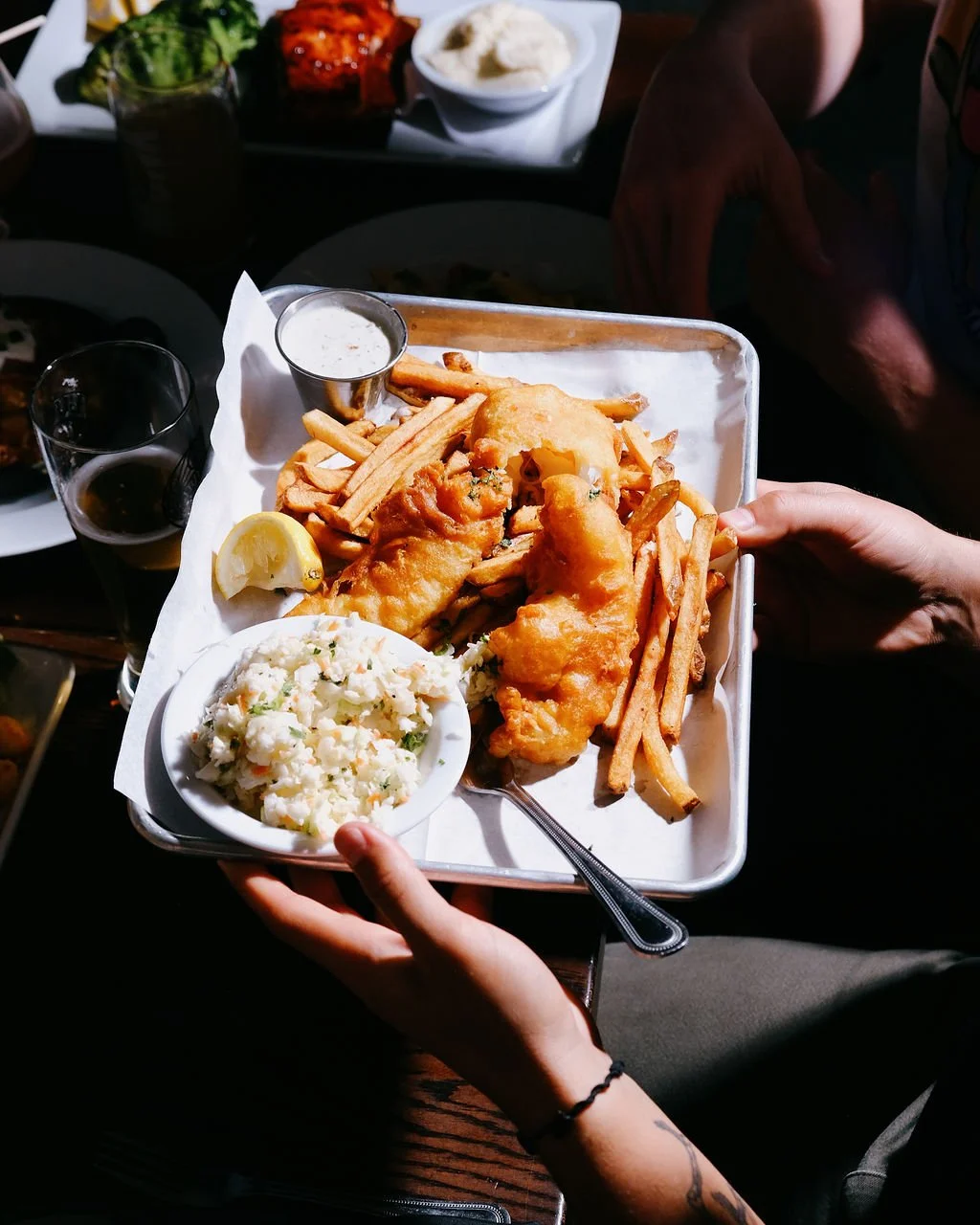 A person holding a tray with fish and chips, French fries, tartar sauce, a lemon wedge, and a side of coleslaw. In the background, there are other plates and drinks on a dark wooden table.
