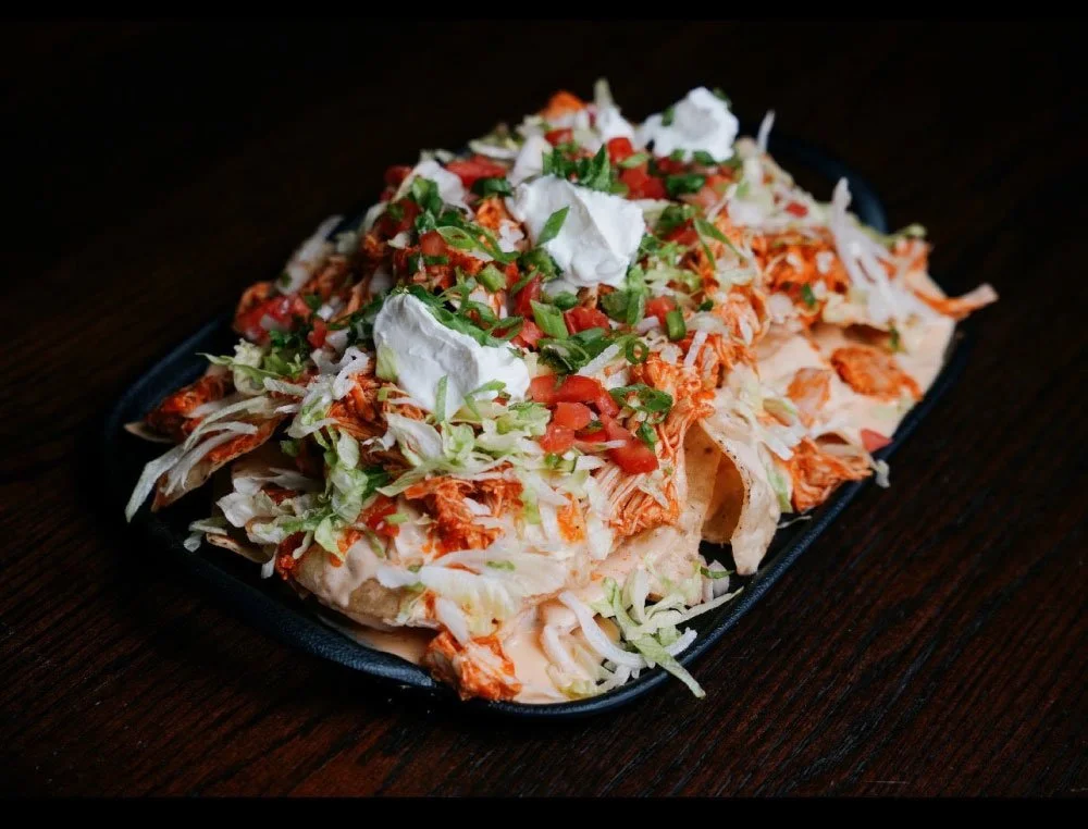 Plate of loaded nachos topped with shredded cheese, chopped tomatoes, green onions, and dollops of sour cream, on a dark wooden table.
