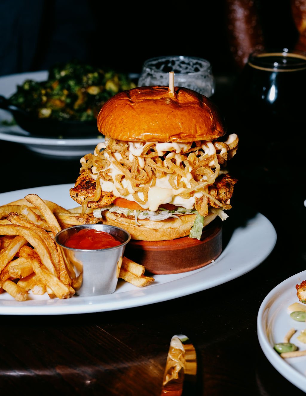 A large burger topped with fried onions on a bun, served with French fries and ketchup in a small metal cup, on a white plate; in the background are glasses of dark beverages and a bowl of salad
