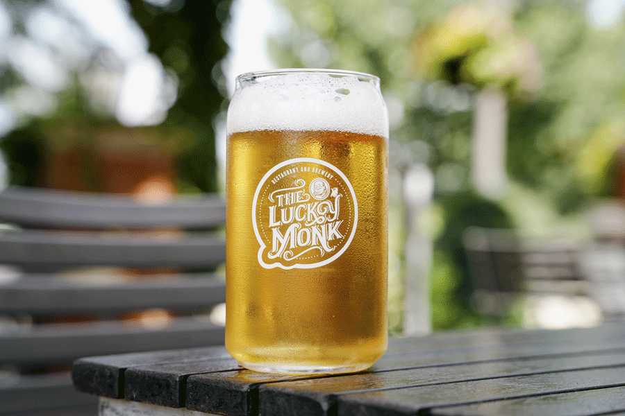 A glass of beer with foam on top, sitting on a wooden table outdoors. The glass has a logo that reads 'Thule Lucky Monk.'