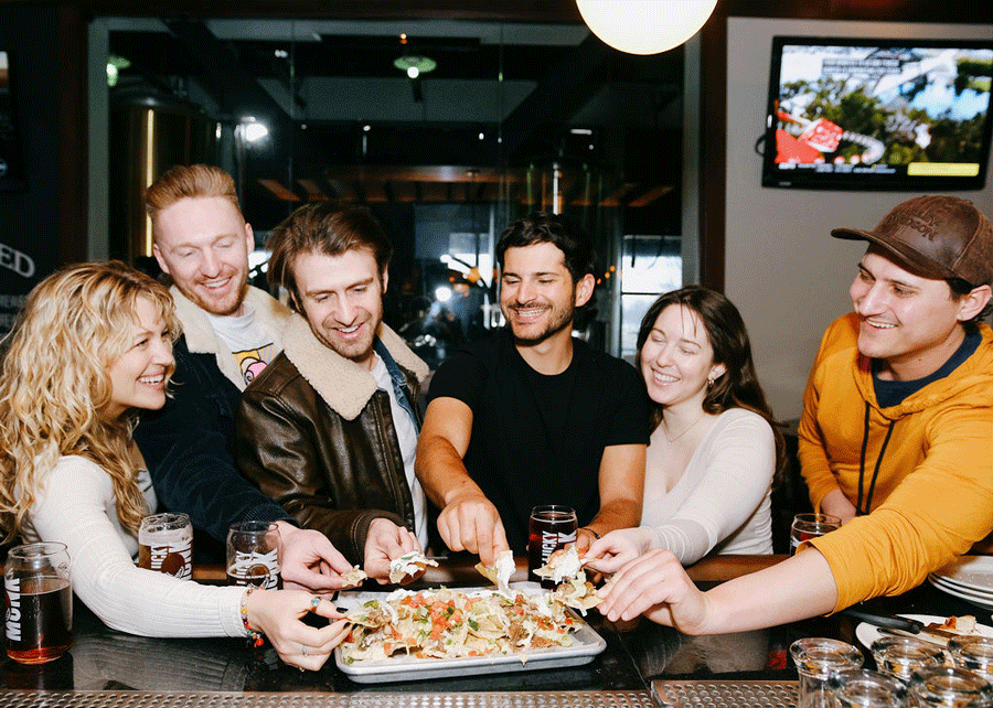 Six young adults sharing a pizza at a bar or restaurant, smiling and enjoying each other's company.