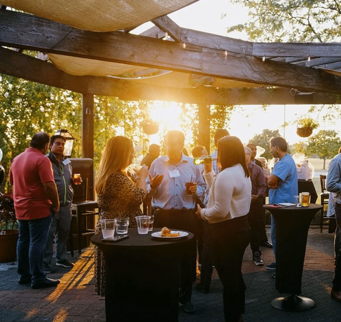 People socializing outdoors at an evening event with string lights and hanging plants, standing around cocktail tables with drinks and snacks.