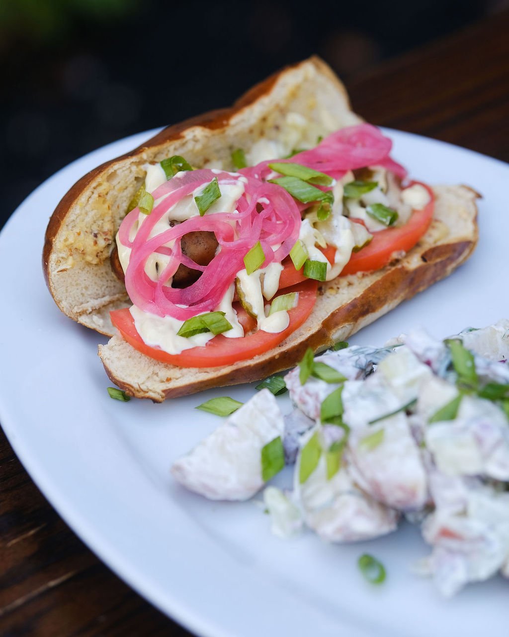 Hot dog bun with tomato slices, pickled onions, mayonnaise, and chopped green onions, served with a side salad.