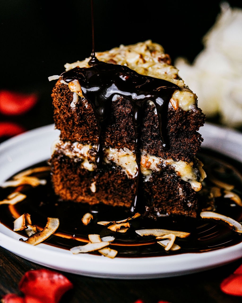 A slice of chocolate cake topped with chocolate syrup and cream, served on a plate with coconut flakes and surrounded by strawberries.