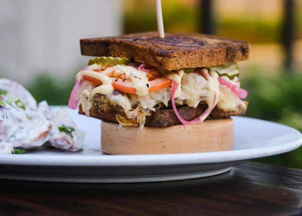 Sandwich with pulled meat, pickles, coleslaw, and mayonnaise, served on toasted bread, with a side of potato salad.
