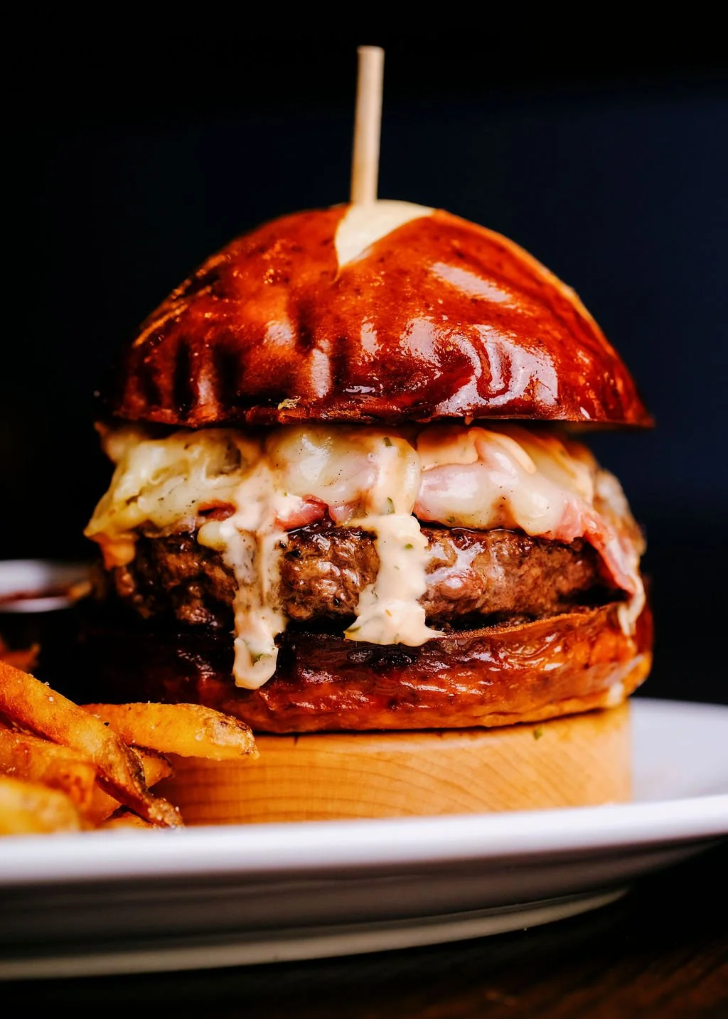 A gourmet burger with melted cheese, a beef patty, a glazed bun, and a side of French fries on a white plate.