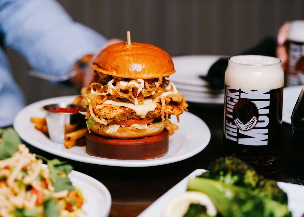 A gourmet burger with fried onion strings, cheese, lettuce, tomato, and sauce on a shiny bun, served with fries and a small cup of ketchup, alongside a glass of dark beer with foam, on a black table with other plates of food and drinks in the background.
