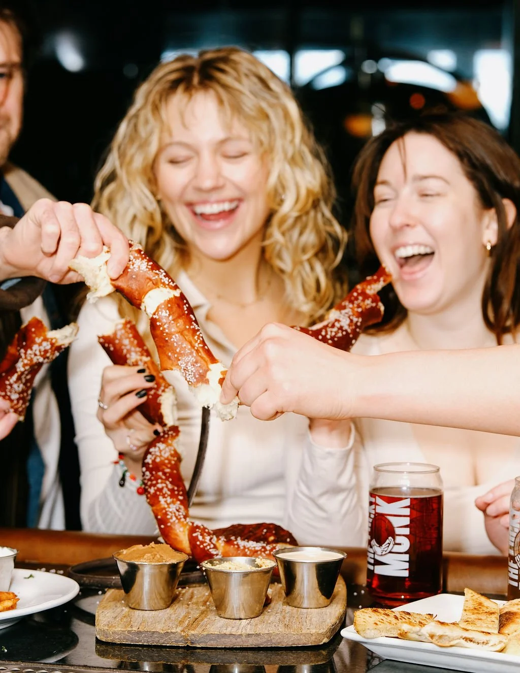 People sharing a large pretzel at a restaurant, smiling and enjoying food and drinks.