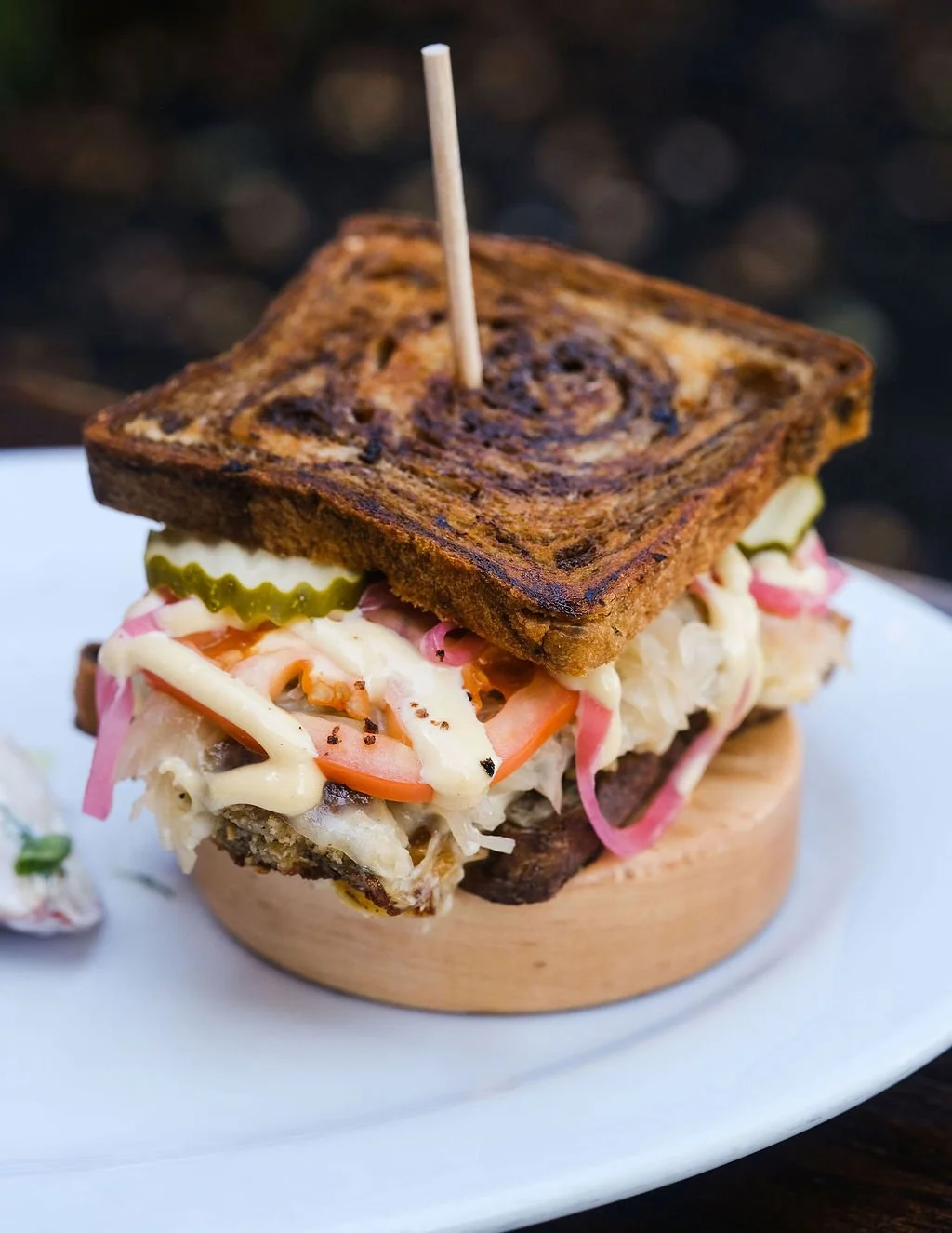 A grilled sandwich with coleslaw, pickles, and vegetables on a wooden stand, topped with a toasted bread slice and a toothpick.