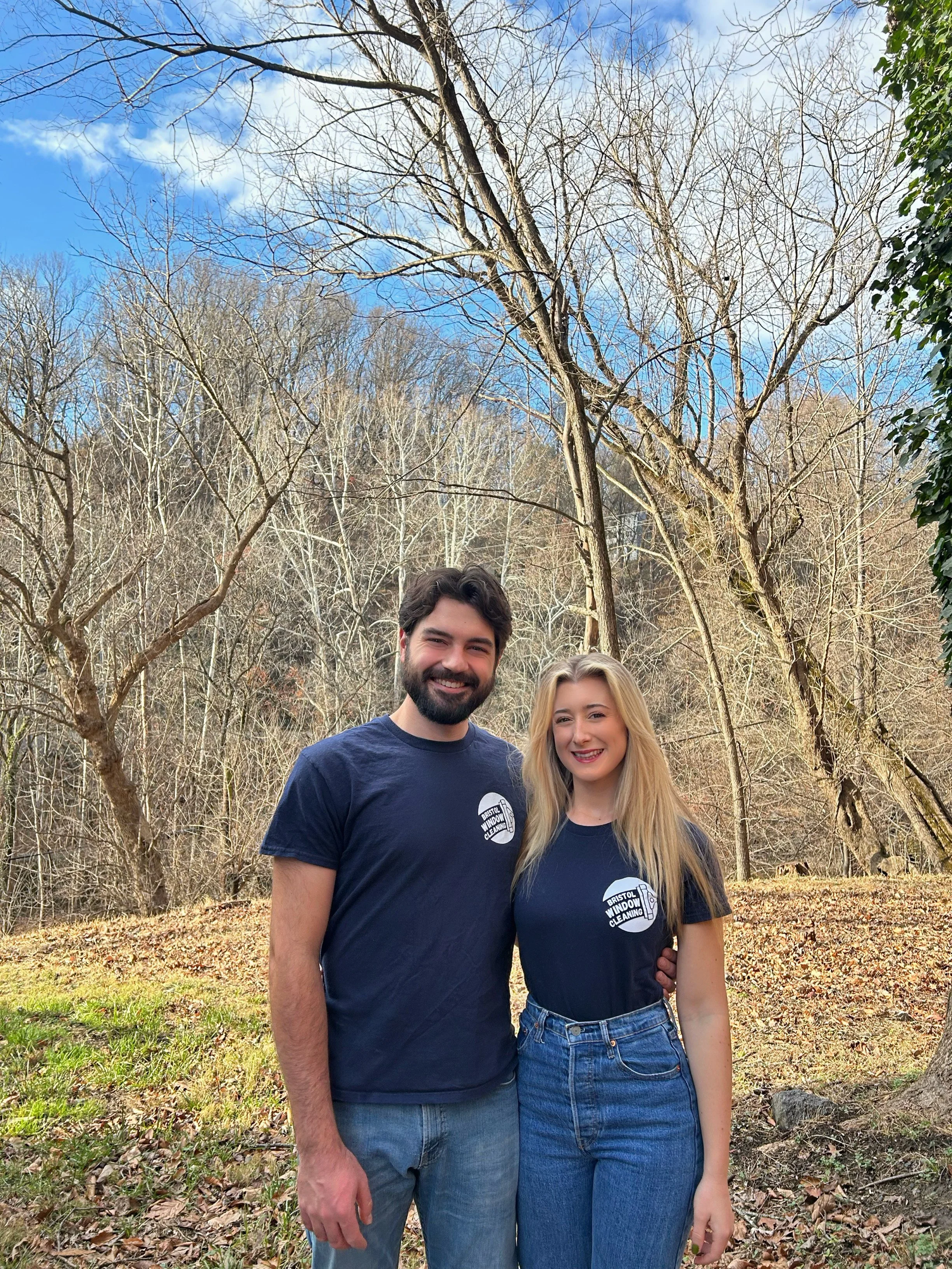 Two people standing outdoors in a wooded area, wearing matching dark t-shirts with the Bristol Window Cleaning logo, smiling for the camera.