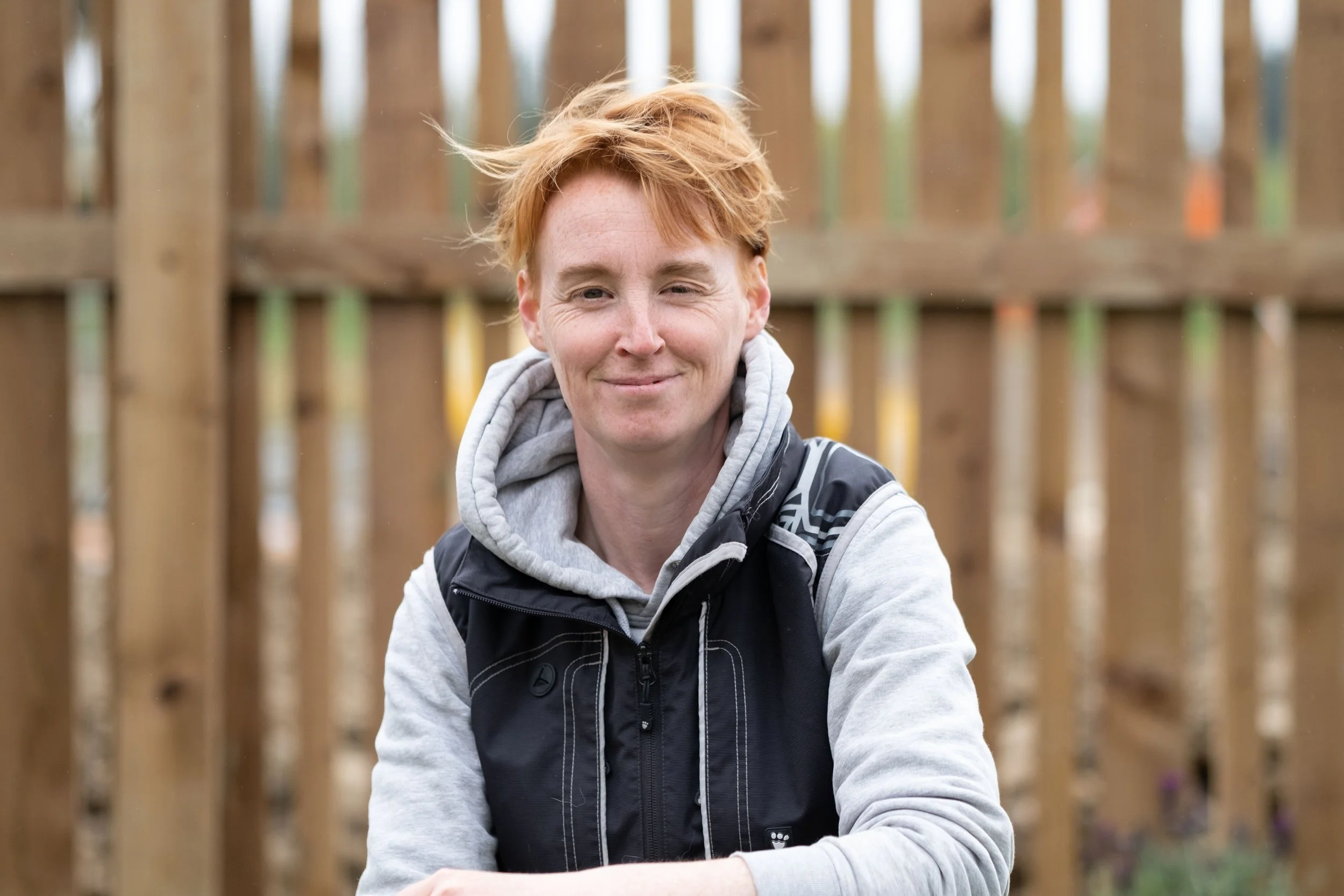 A woman with short red hair smiling outdoors, sitting in front of a wooden fence, wearing a gray hoodie and black vest.