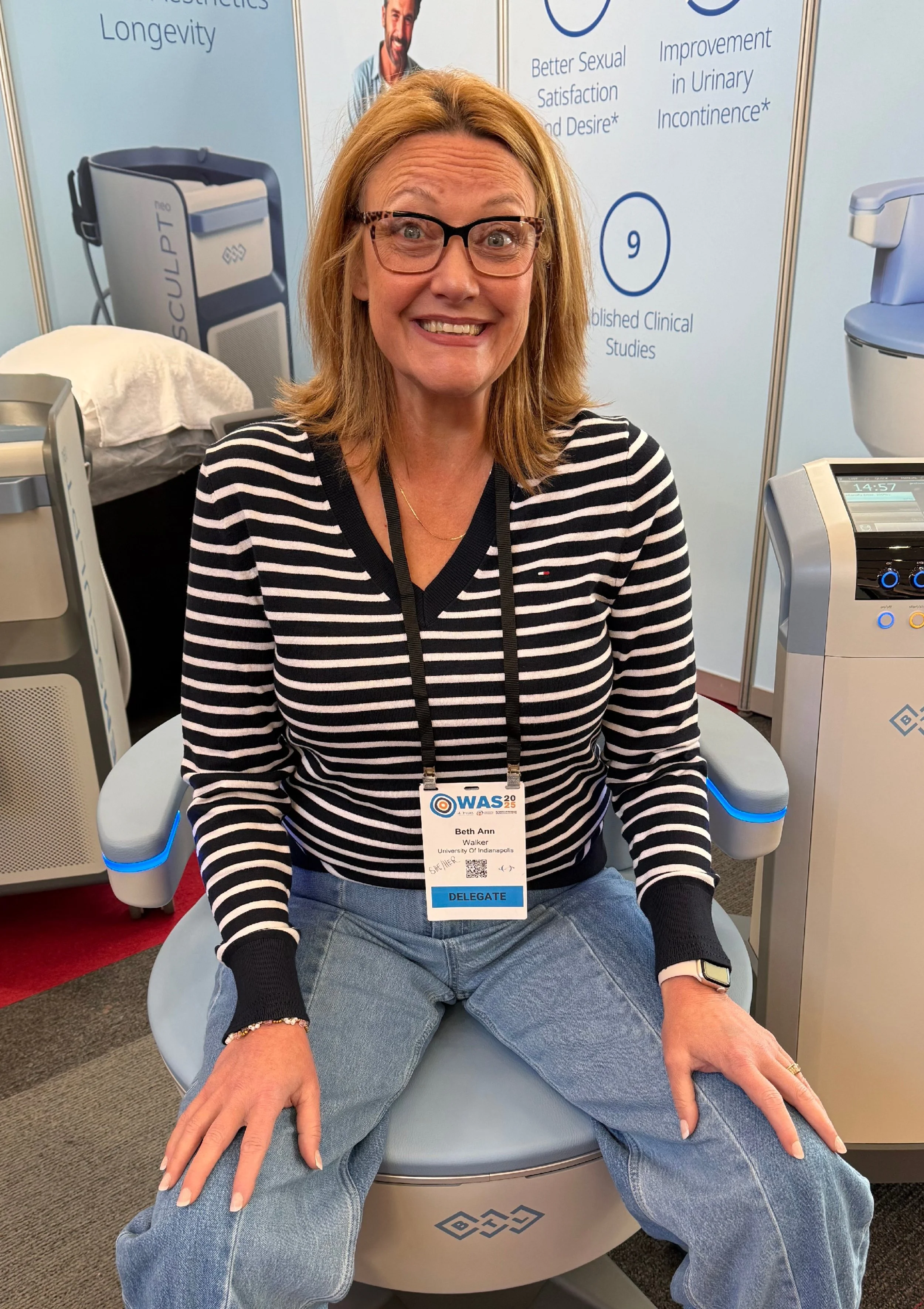 Woman with glasses and a name badge sitting in a medical device, smiling at the camera. Background contains medical imaging equipment and informational posters about clinical studies.