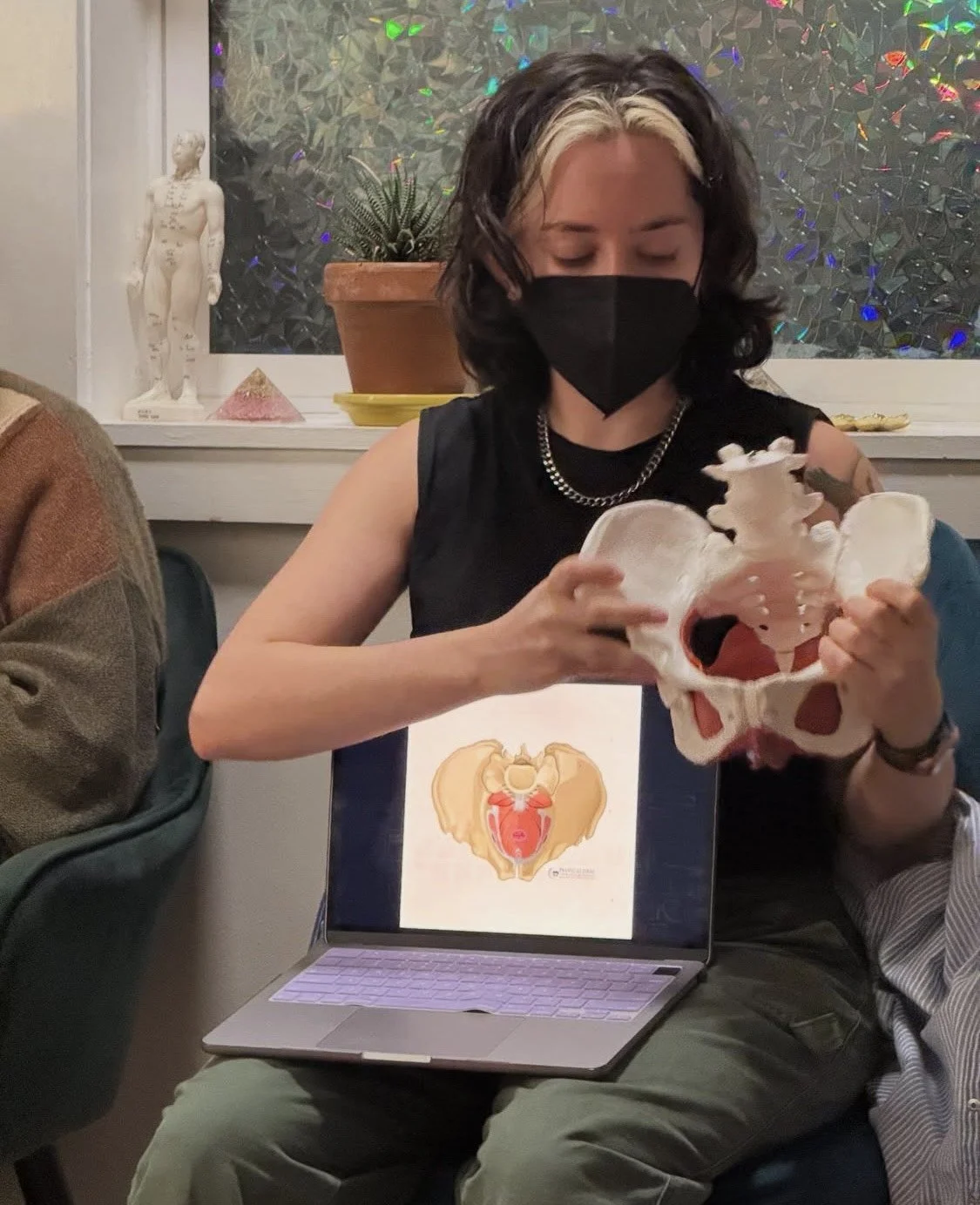 Person with dark hair and a black face mask sitting at a table holding a large anatomical model of a pelvis and reproductive system, with a laptop displaying an illustration of the same anatomy, in a room with window decorations and a potted plant.