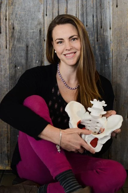 A woman smiling and holding a model of a human pelvis, sitting against a wooden background.