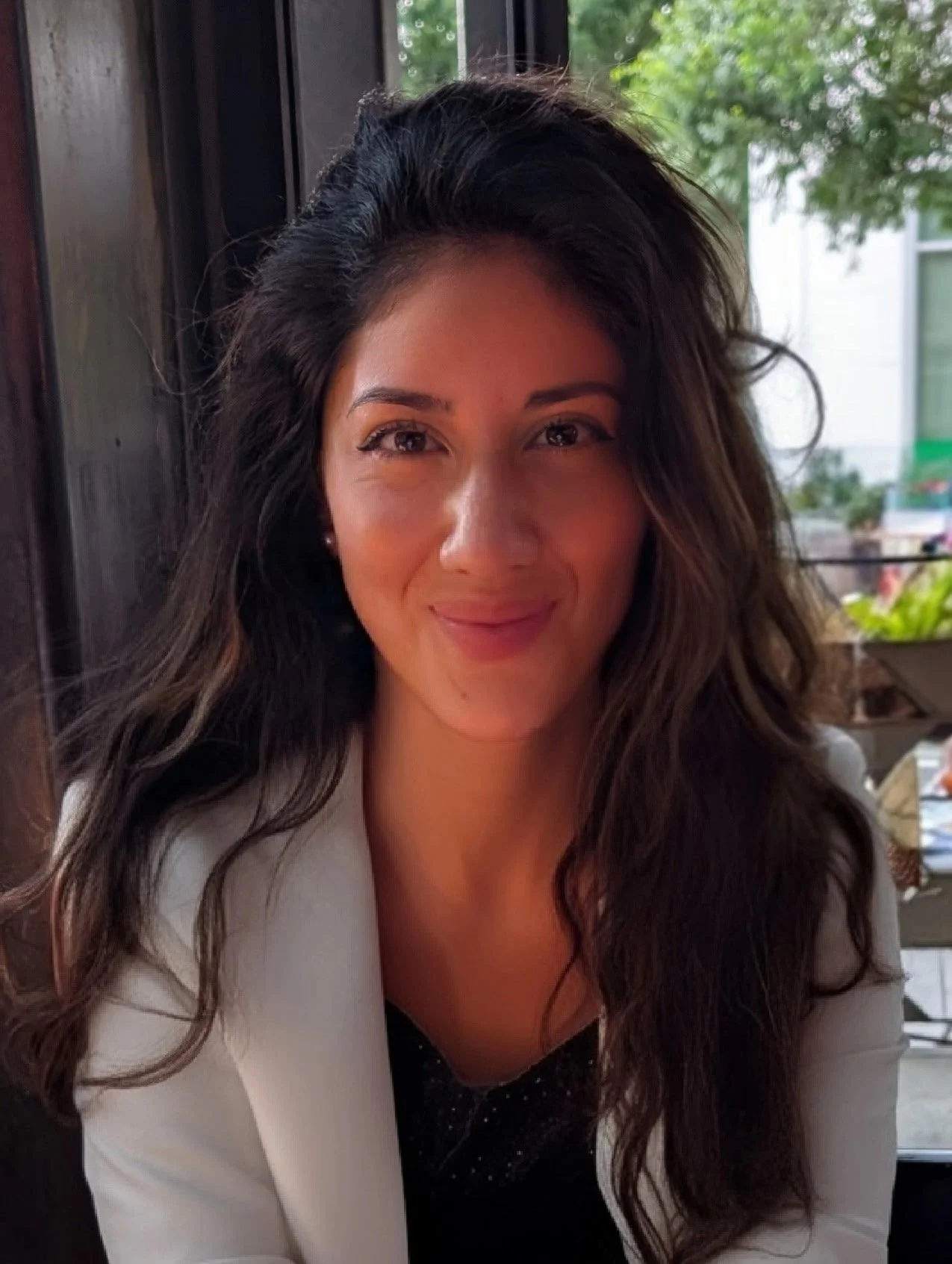 A young woman with long wavy dark hair, smiling softly, sitting indoors near a window with greenery outside.