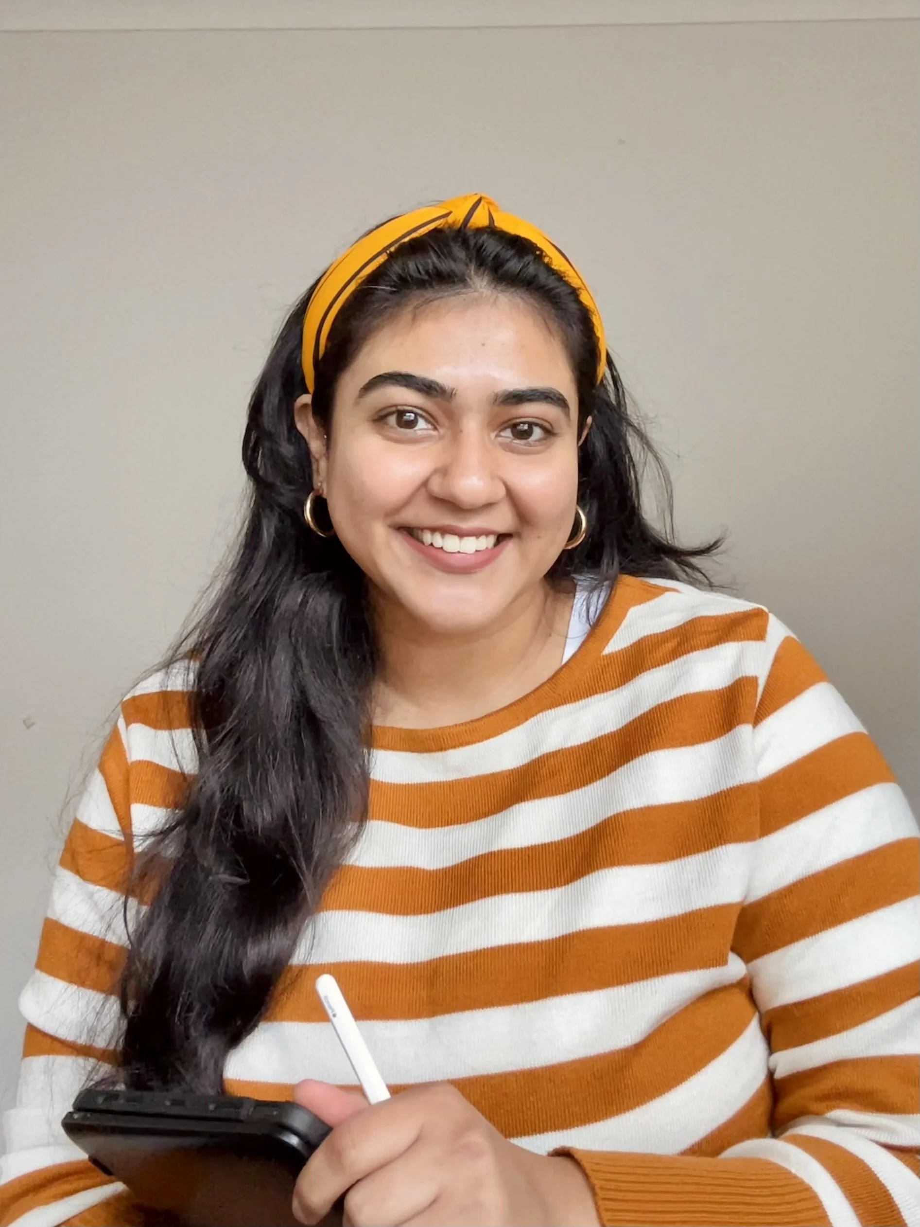 A young woman with long dark hair, wearing a yellow headband and a striped orange and white sweater, smiling while holding a smartphone and stylus in her hands.