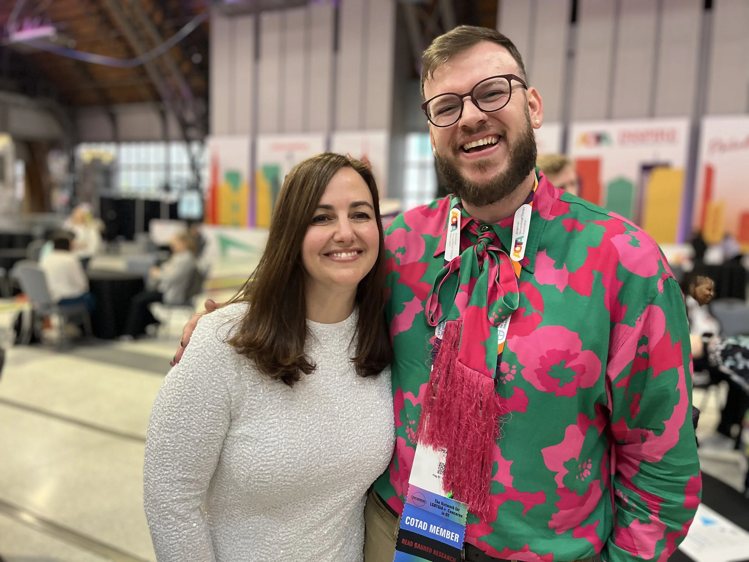 Two people smiling and posing at a conference. The woman has shoulder-length brown hair and is wearing a white textured top. The man has short brown hair, glasses, a beard, and is wearing a bright pink and green floral shirt with a pink fringed scarf. They are standing indoors with a blurred background of conference tables and people.