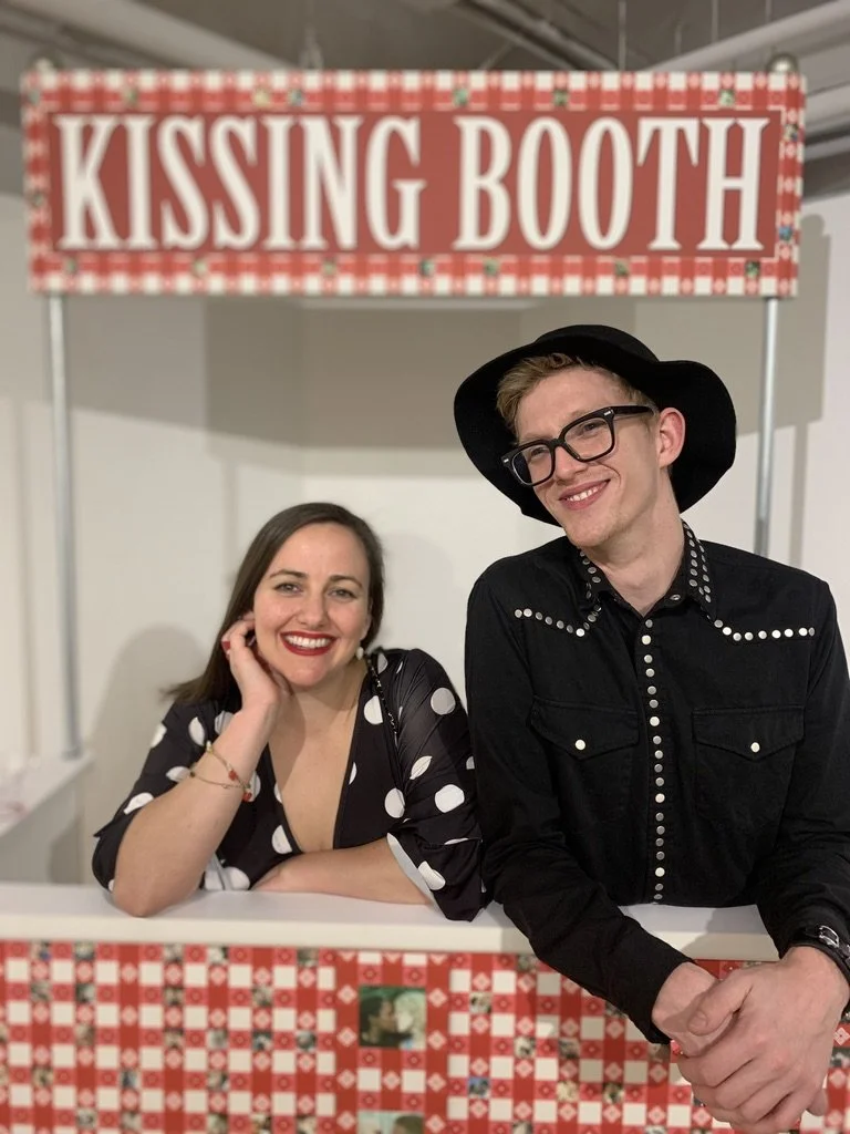 A smiling woman with long brown hair and a woman with short blond hair wearing glasses and a black hat, standing behind a kissing booth with a pink and red checkered and floral border and sign that reads "Kissing Booth."