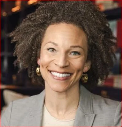 A woman with curly hair smiling, wearing earrings and a blazer, indoors with bookshelves in the background.