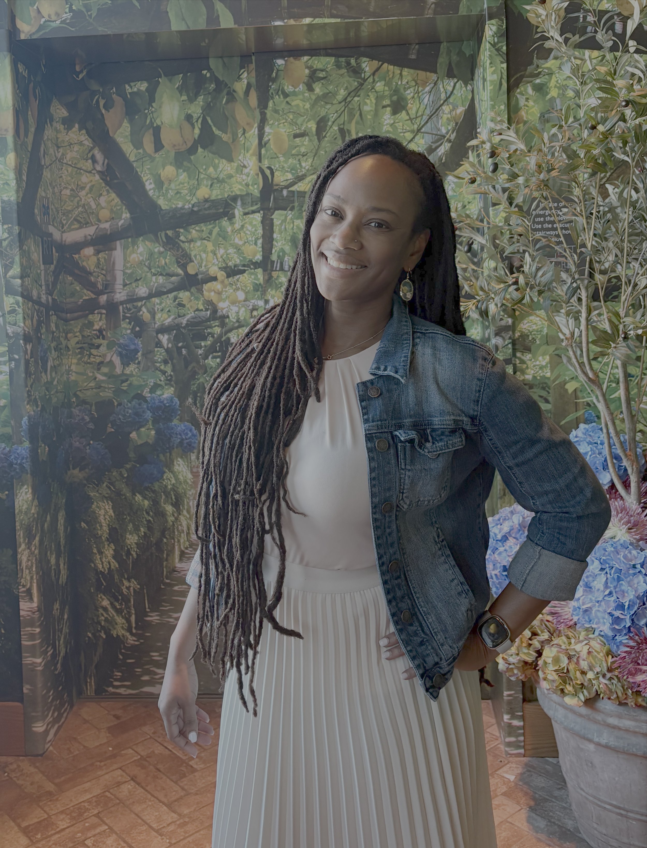 A woman with long dreadlocks and wearing a denim jacket and a white pleated skirt, standing indoors with a leafy, green backdrop and a pot with yellow flowers beside her.