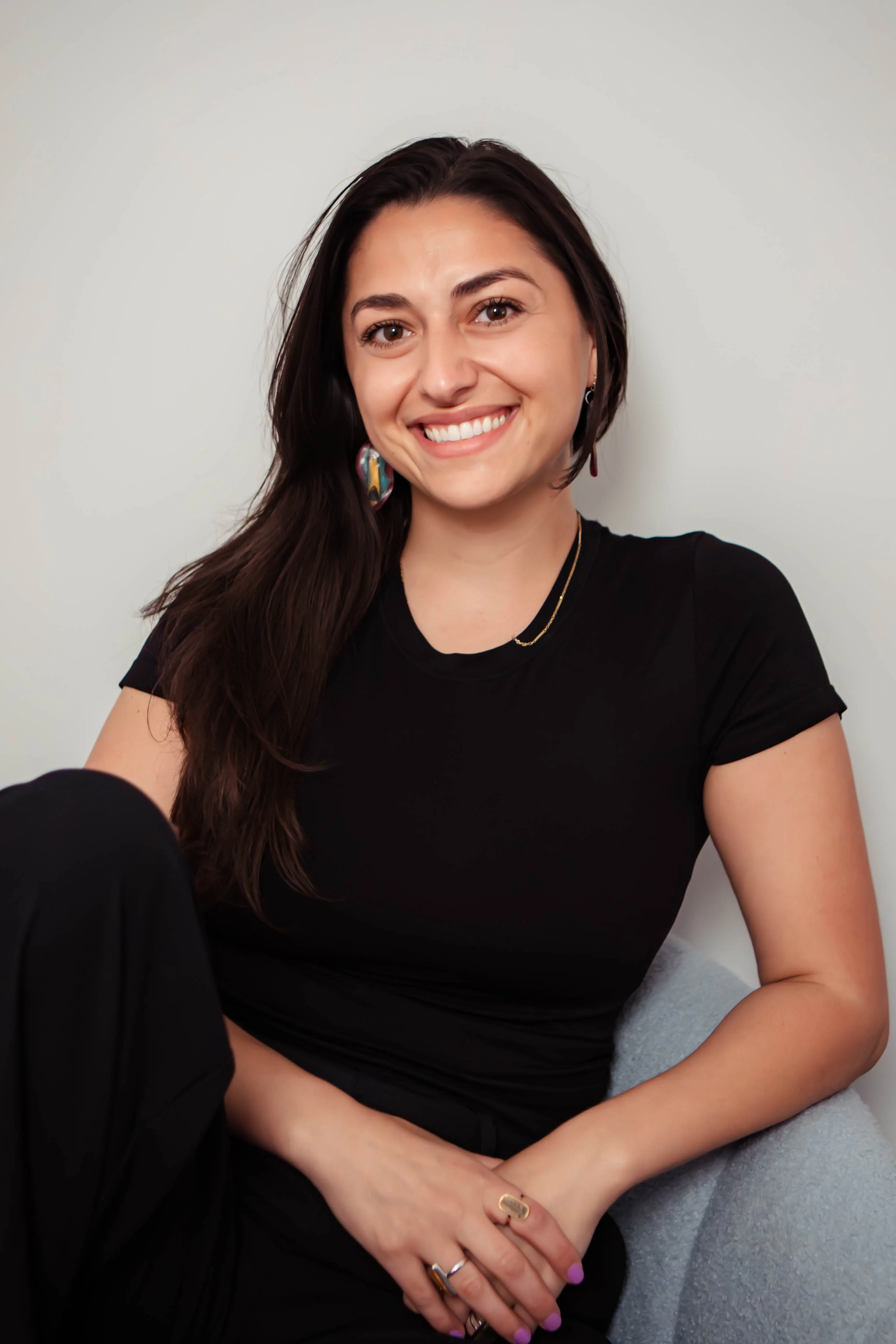 A woman with long dark hair, smiling, wearing a black t-shirt, colorful earrings, a gold necklace, and rings, sitting against a plain light-colored wall.
