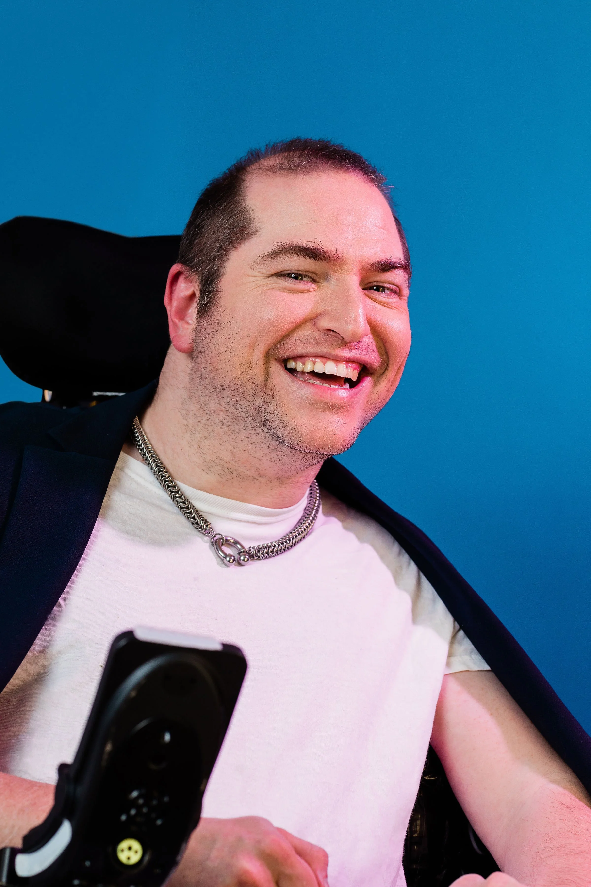 A man smiling and looking at a camera, with a blue background, wearing a white T-shirt, a silver chain necklace, and a black jacket, with a wheelchair visible behind him.