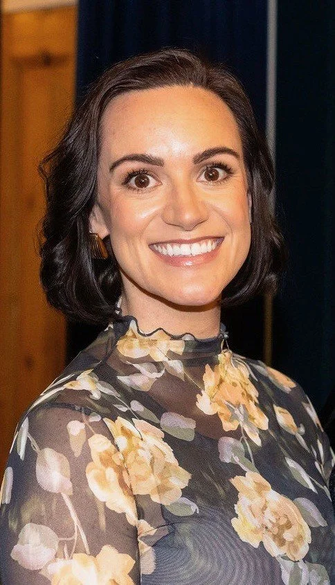 A woman with short dark hair smiling, wearing a floral-patterned top, standing indoors with a wooden wall and dark curtains in the background.