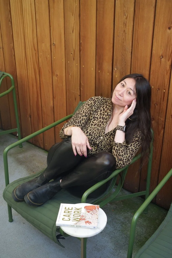 A woman with long dark hair wearing a leopard print top, black leather pants, and black ankle boots, sitting on a green outdoor chair with her legs crossed. She is resting her head on her left hand and looking at the camera. A book titled 'CARE CARE' is on a small round table next to her, with a wooden fence in the background.