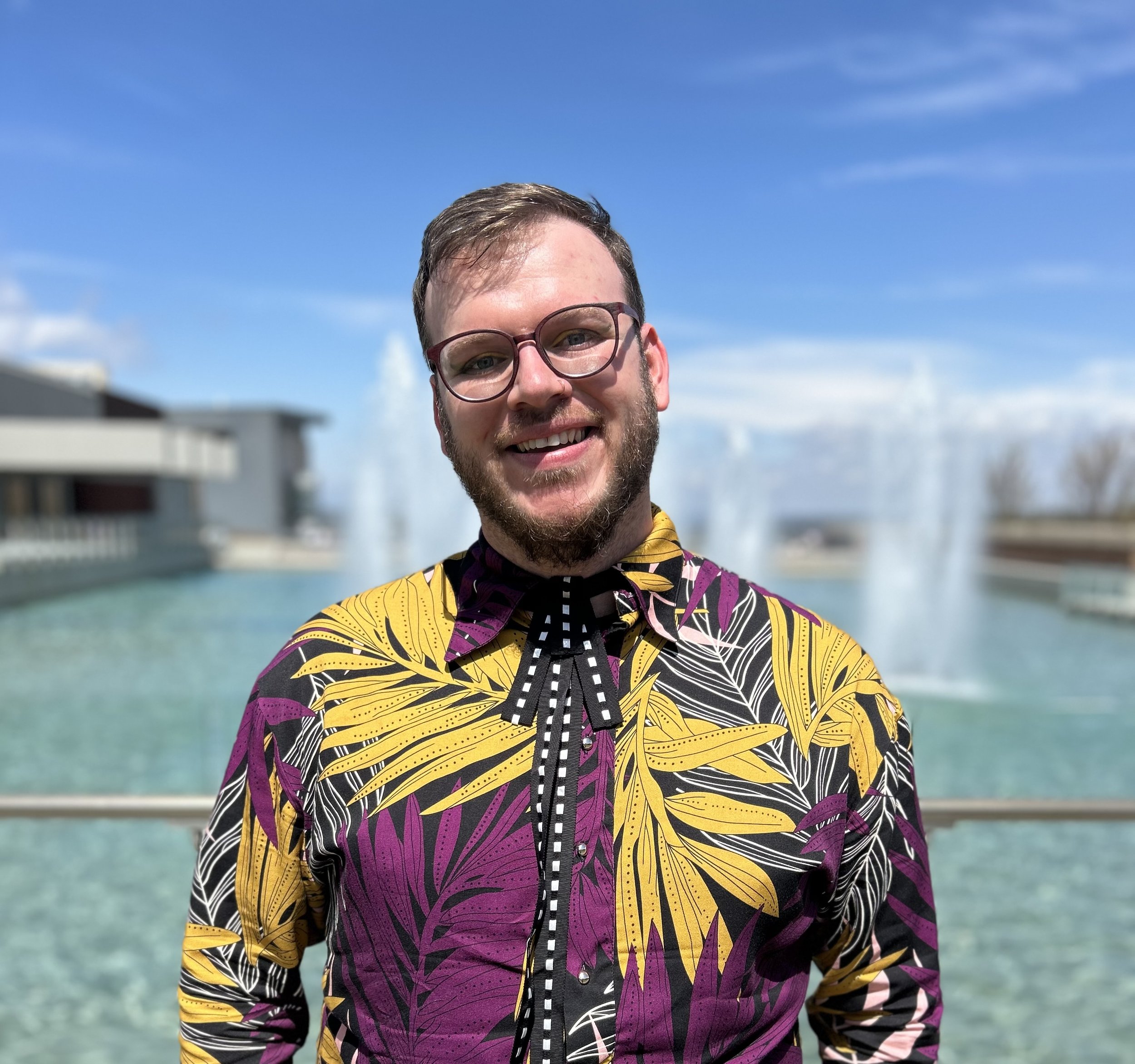 Smiling man with glasses and a beard standing outdoors in front of a water fountain and modern buildings under a blue sky.