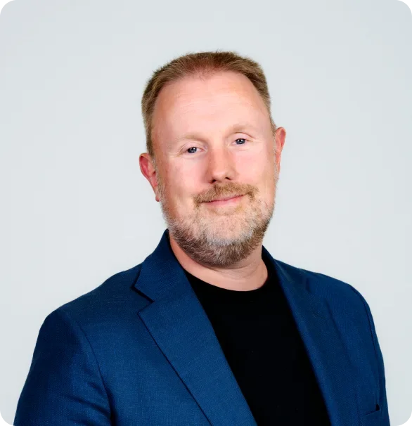 Portrait of a middle-aged man with a beard and short hair, wearing a dark blue blazer and a black shirt, smiling against a light gray background.