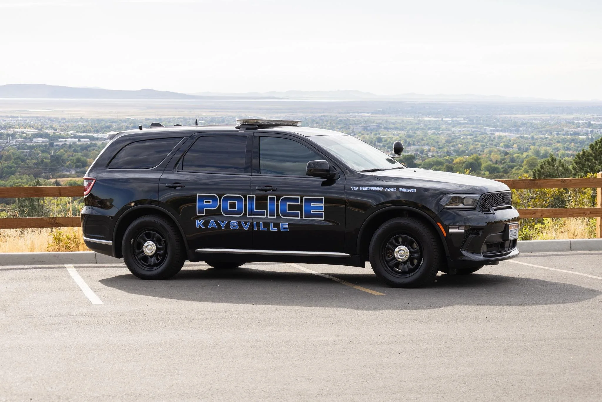 Black police SUV with 'POLICE KAYSVILLE' markings parked in a parking lot on a hill overlooking a city landscape with trees and mountains in the background.