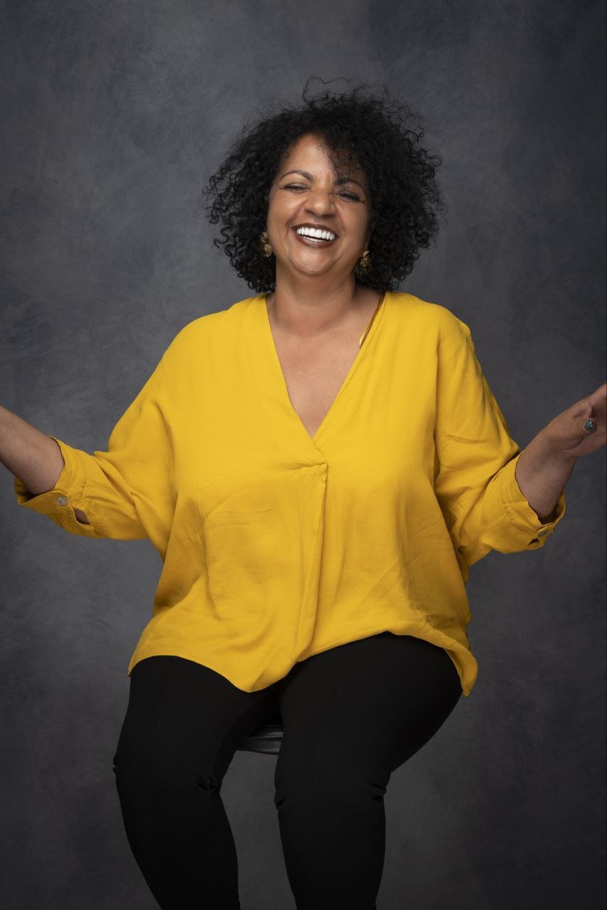 Eine lachende Frau mit lockigem schwarzen Haar, die eine gelbe Bluse trägt, sitzt vor einem grauen Hintergrund.