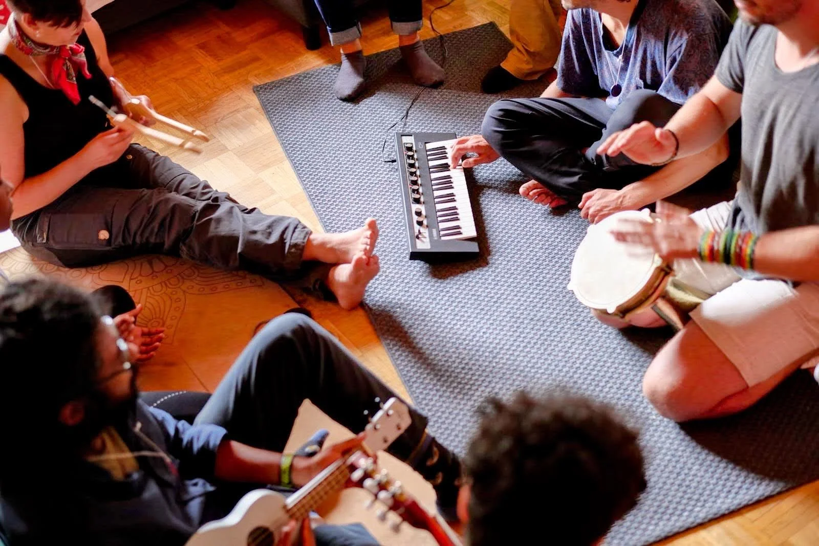 Gruppenspieler sitzen im Kreis auf dem Boden, spielen Musikinstrumente wie Gitarre, Tamburin und ein Keyboard, in einem Raum mit Holzboden und Teppich.