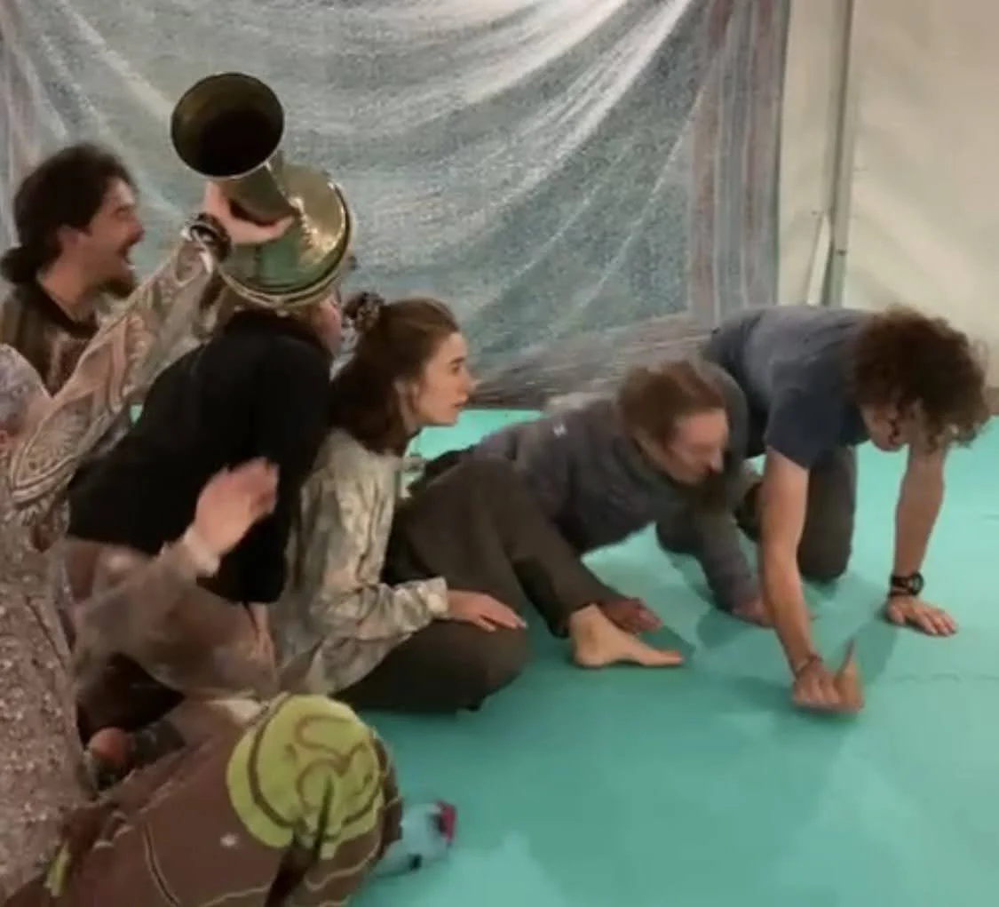 Drei Frauen und eine Person in einer Zirkusaktivität, die auf den Händen und Knien auf einer grünen Matte auf dem Boden krabbeln, während eine andere Frau sie beobachtet und eine Person im Hintergrund einen Pokal hochhält.