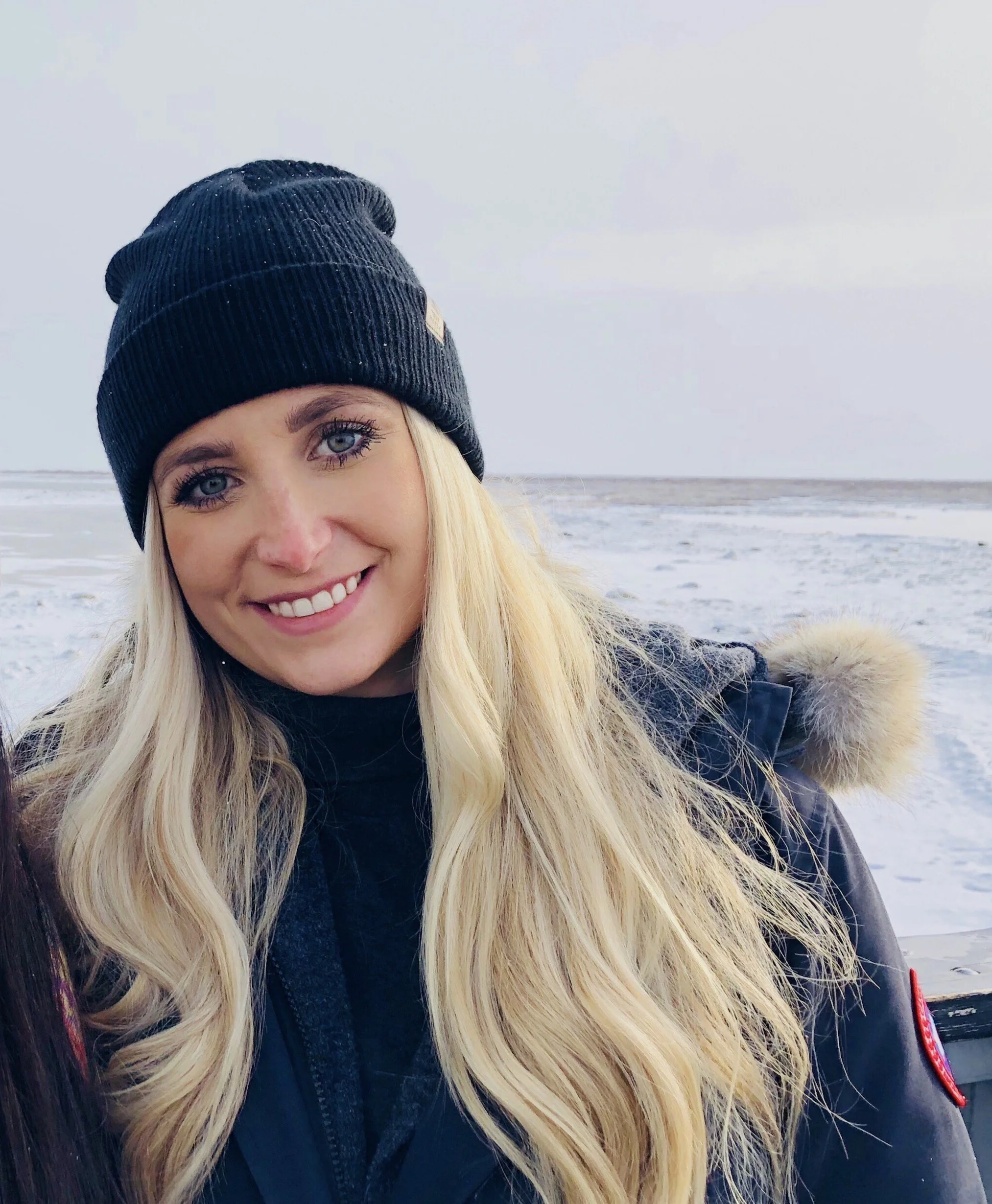 Woman in winter clothing with long blonde hair and a black beanie, smiling outdoors by a beach.