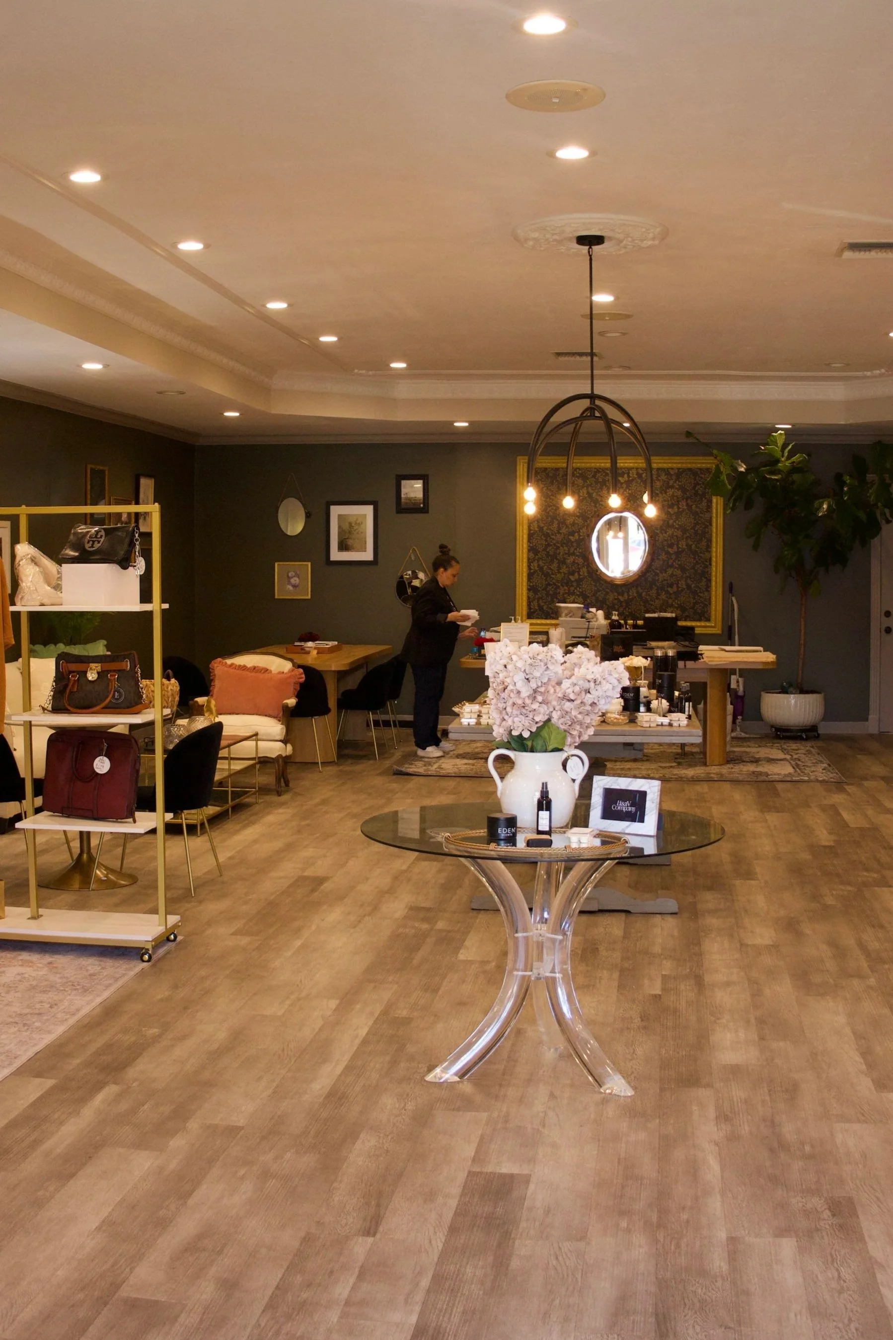 Interior of a boutique with a round glass table holding a white vase filled with light pink flowers, against a wooden floor. Shelves with handbags to the left and a person arranging items at a counter in the background. Modern ceiling lights and wall decorations.